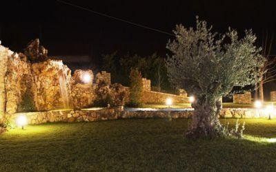 Giardino notturno con muri in pietra illuminati, prato e un grande albero sotto luci calde