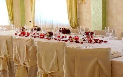 Elegante tavolo da banchetto apparecchiato con tovaglie bianche, dettagli rossi e calici da vino in una luminosa sala da pranzo.