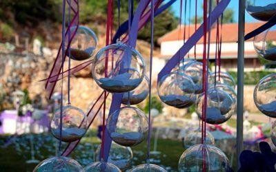 Campanelli a vento in vetro appesi all'aperto in un giardino soleggiato, con alberi e fiori sfocati sullo sfondo.