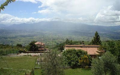 Valle verdeggiante con case dai tetti di tegole, alberi e montagne in lontananza sotto un cielo nuvoloso.