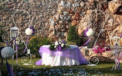 Tavolo nuziale viola allestito all'aperto accanto a un muro di pietra con decorazioni floreali bianche e lanterne.