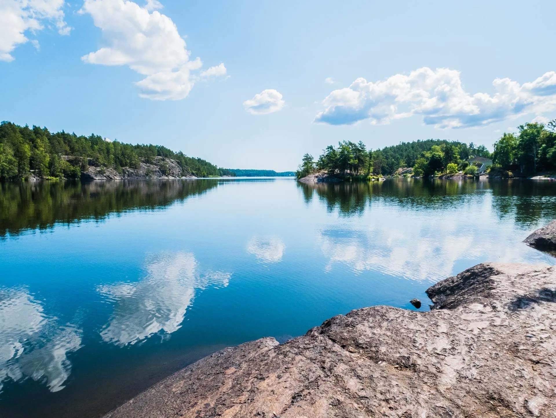 Calm lake reflecting blue skies and clouds, bordered by forest and a rocky shoreline.