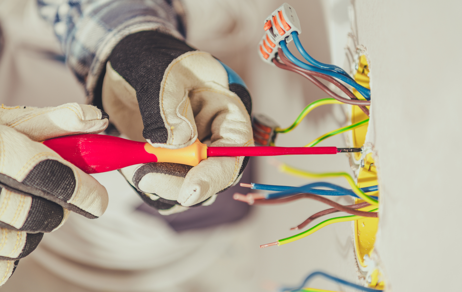 Gloved hands using a red screwdriver to work on colorful electrical wires in a wall box