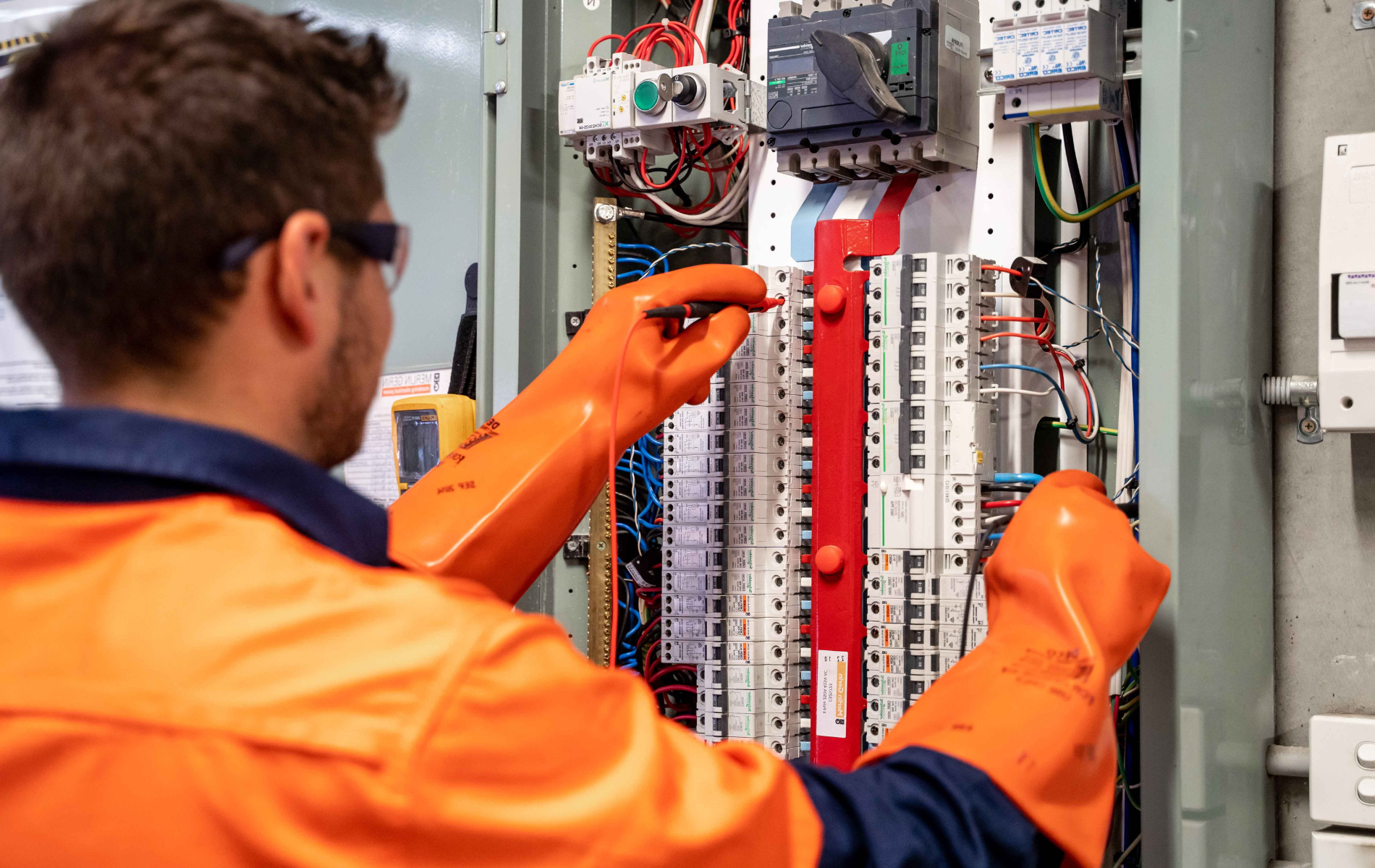 Technician in orange gloves inspecting electrical control panel with wiring and relays