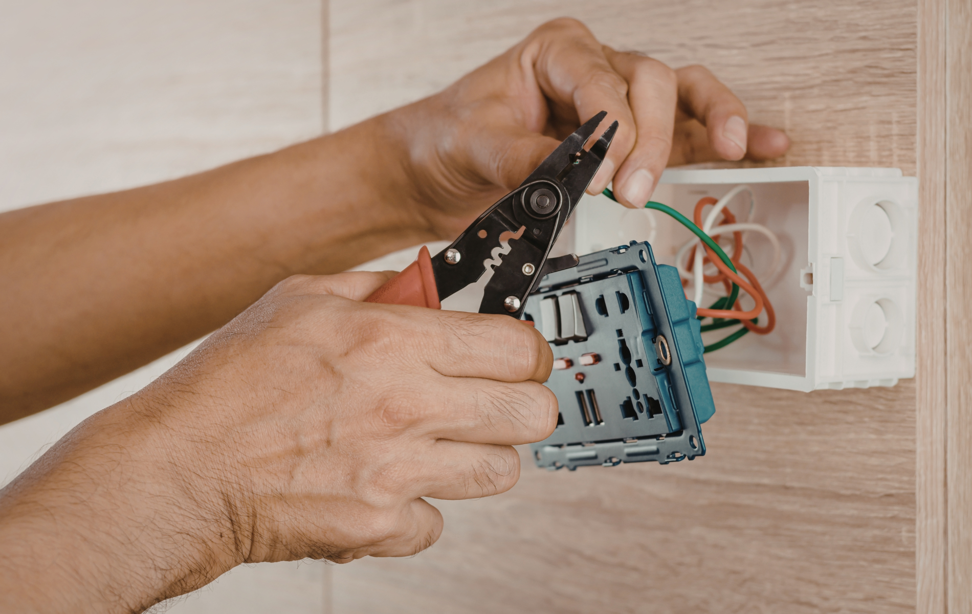 Hands wiring an electrical outlet box in a wall, with exposed cables and a screwdriver