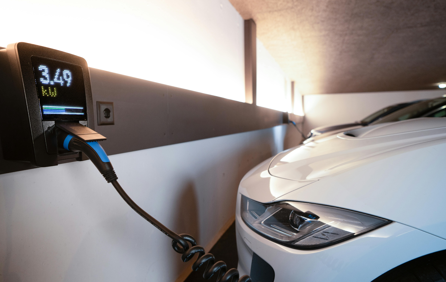Electric car charging in a garage, with a white car plugged into a wall charger showing 3.49.