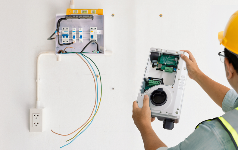 Electrician wiring a wall-mounted electrical panel in a white room, holding a circuit breaker box.