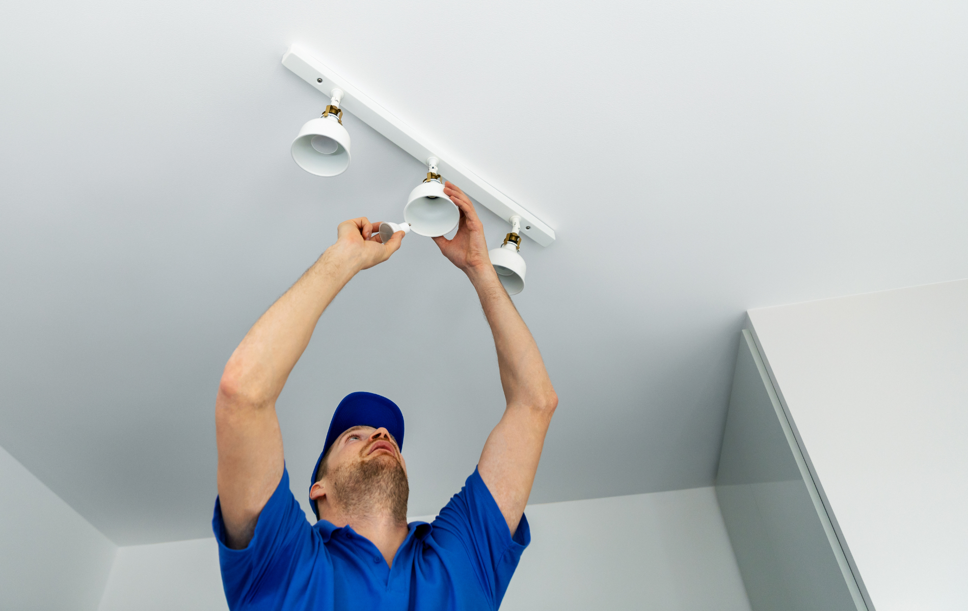 Person replacing a ceiling track light in a white room, reaching up with tools.