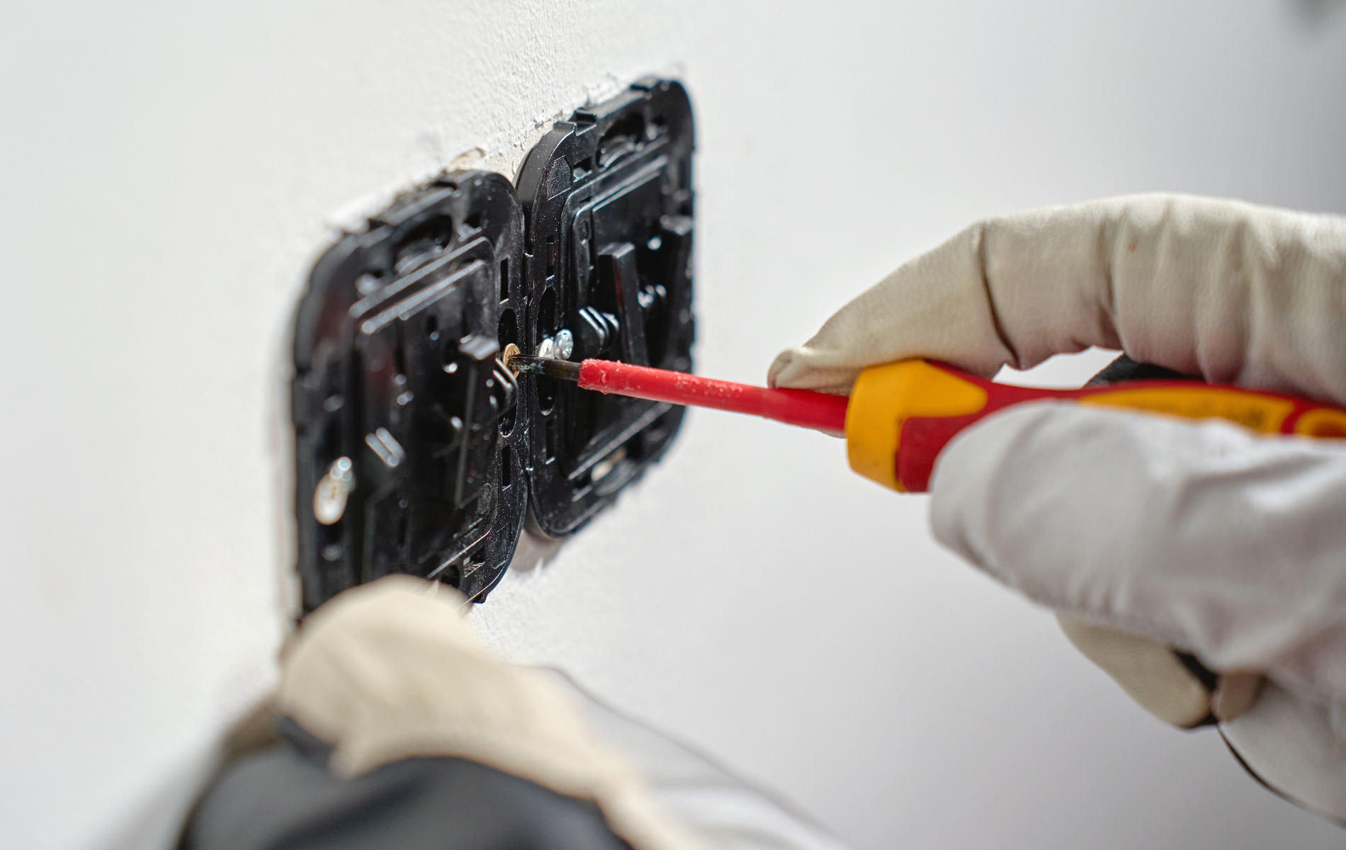 Gloved hands using a screwdriver to work on an electrical outlet on a wall