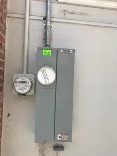 Gray electrical meters and breaker panel mounted on a wall in a utility area.