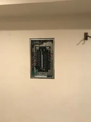 Open electrical breaker panel in a beige wall, with wires visible inside and a small hook nearby.