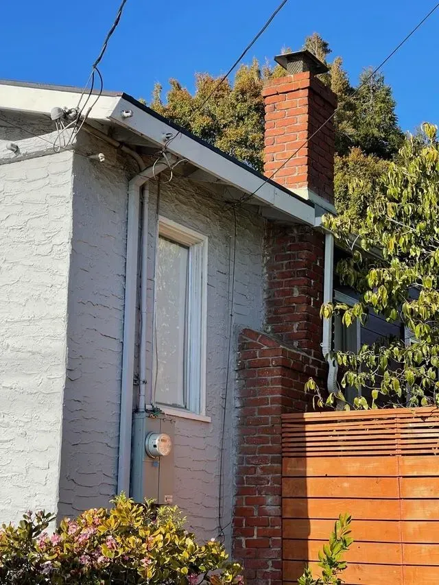 Side of a house with a brick chimney, white window, and wooden fence under a blue sky