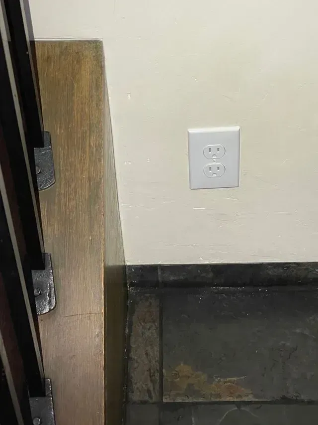White wall with a single electrical outlet above a dark baseboard and a wooden panel on the left.