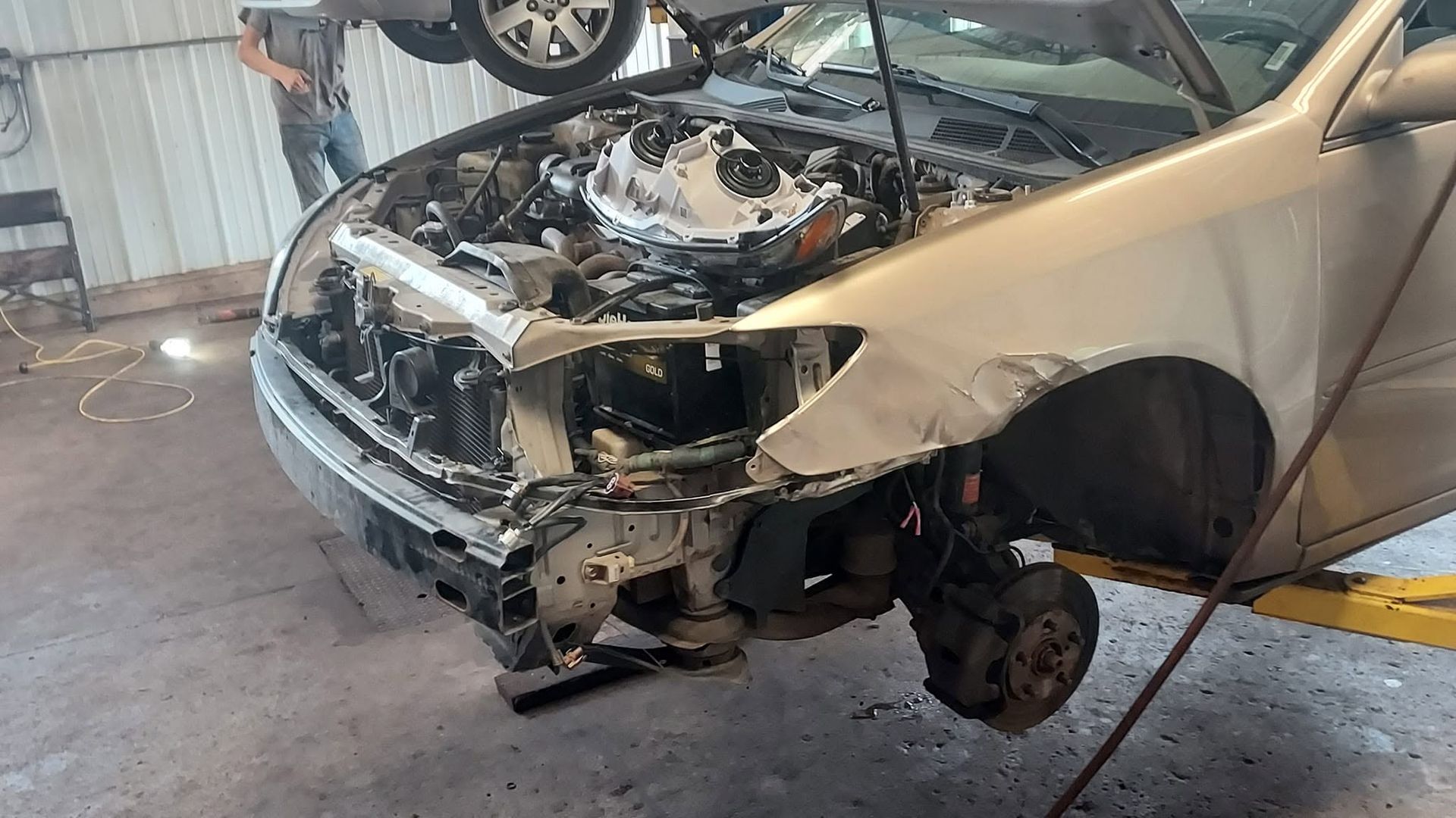 Damaged beige car front end in a repair shop, with the hood open and front bumper removed