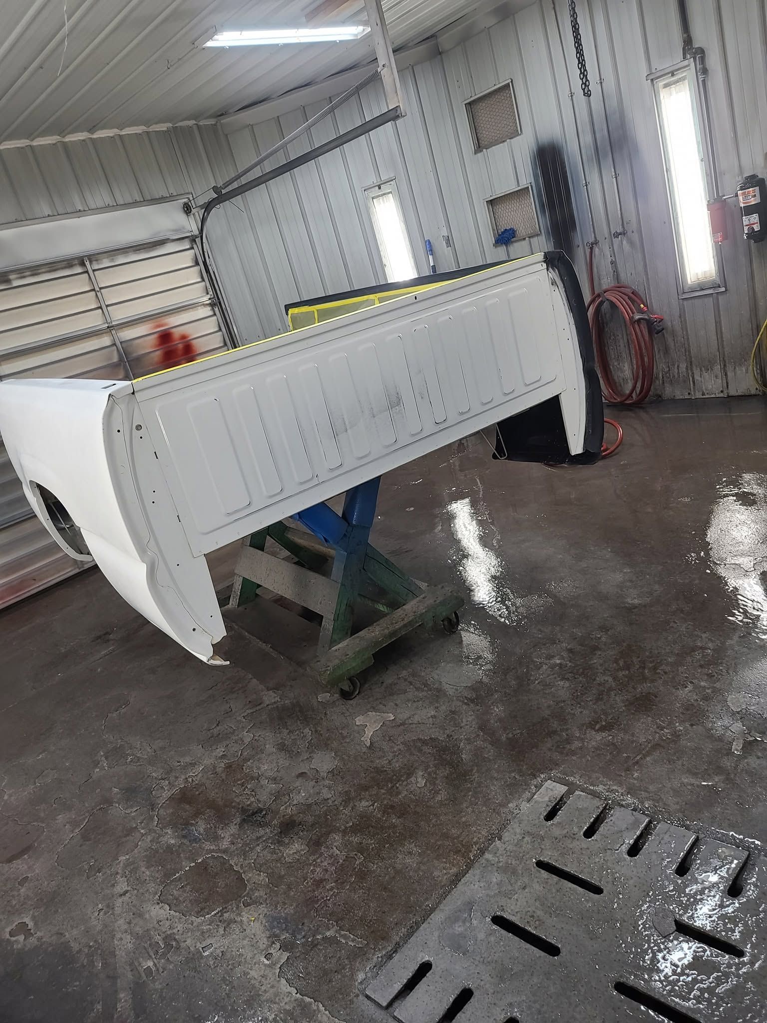 White truck bed in auto body shop on green stand, with wet floor and work lights nearby