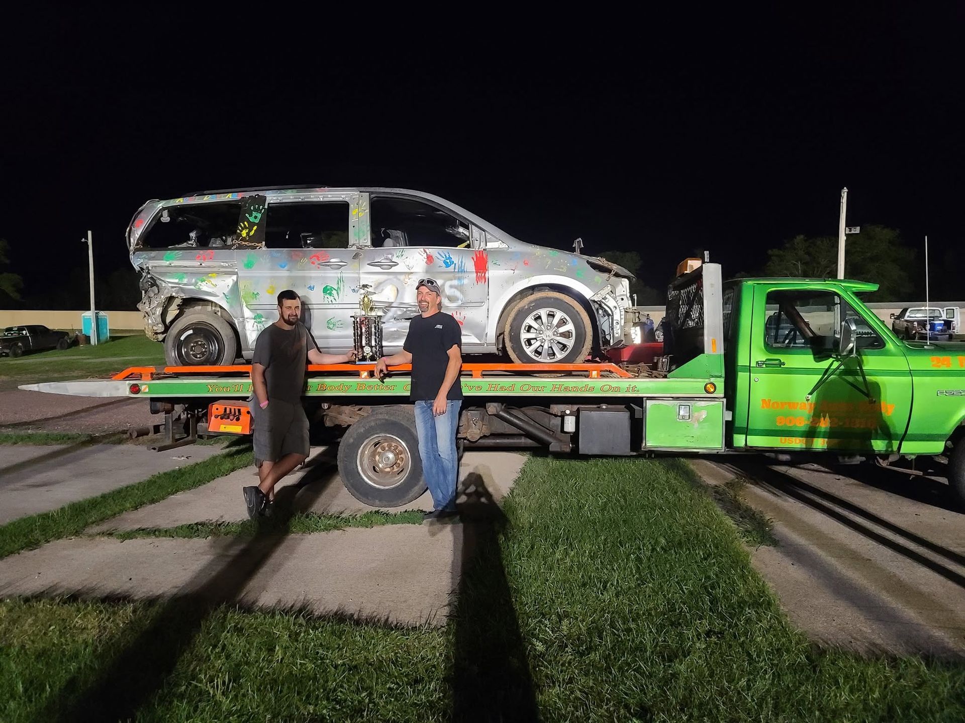 Green tow truck lifting a white van at night, with two people standing beside it.