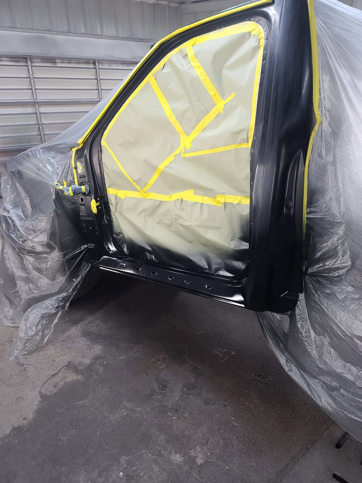Masked car door panel in a workshop, covered in black and yellow tape with plastic sheeting nearby
