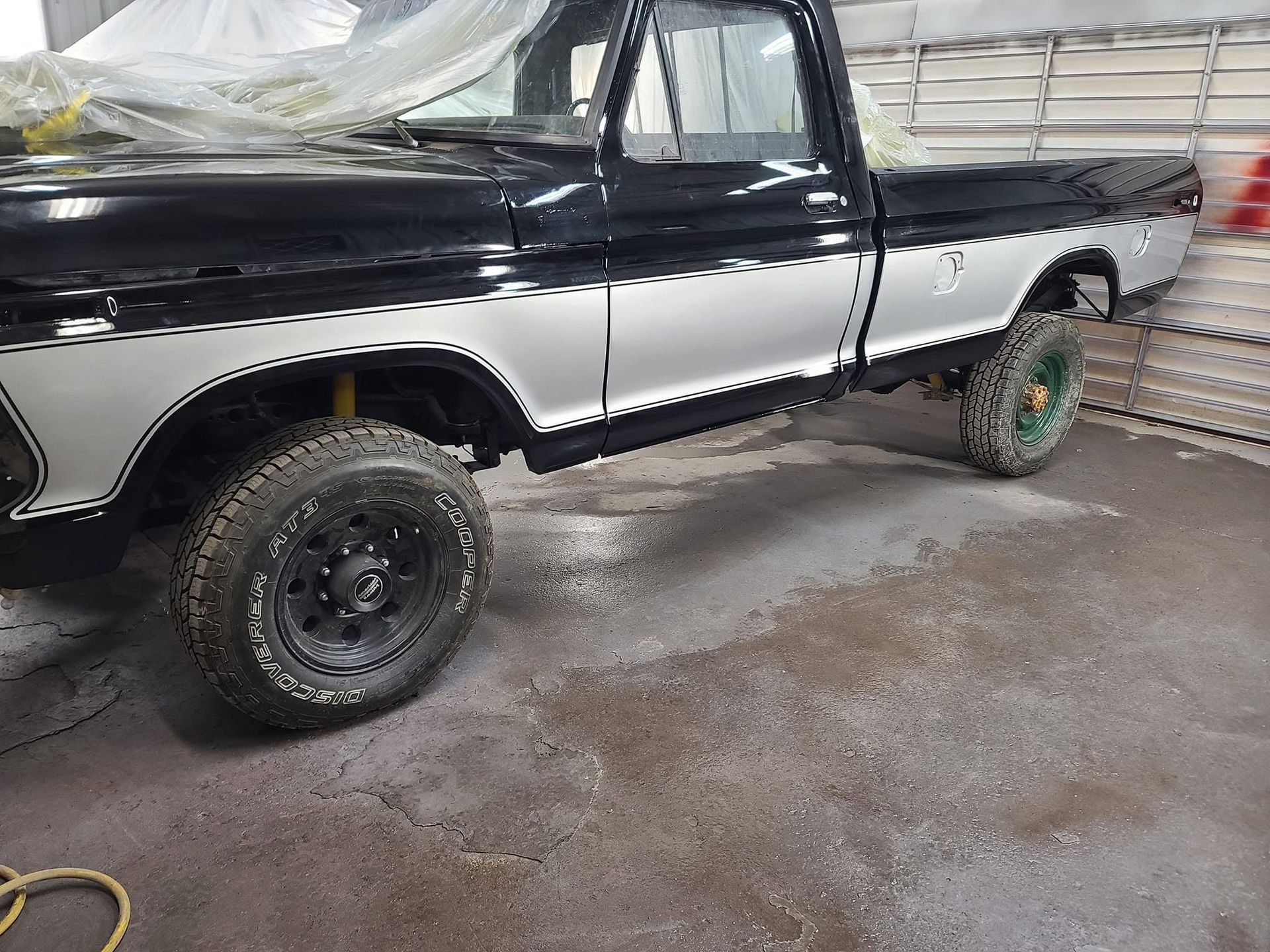 Black-and-white pickup truck with off-road tires parked in a garage beside a corrugated wall.