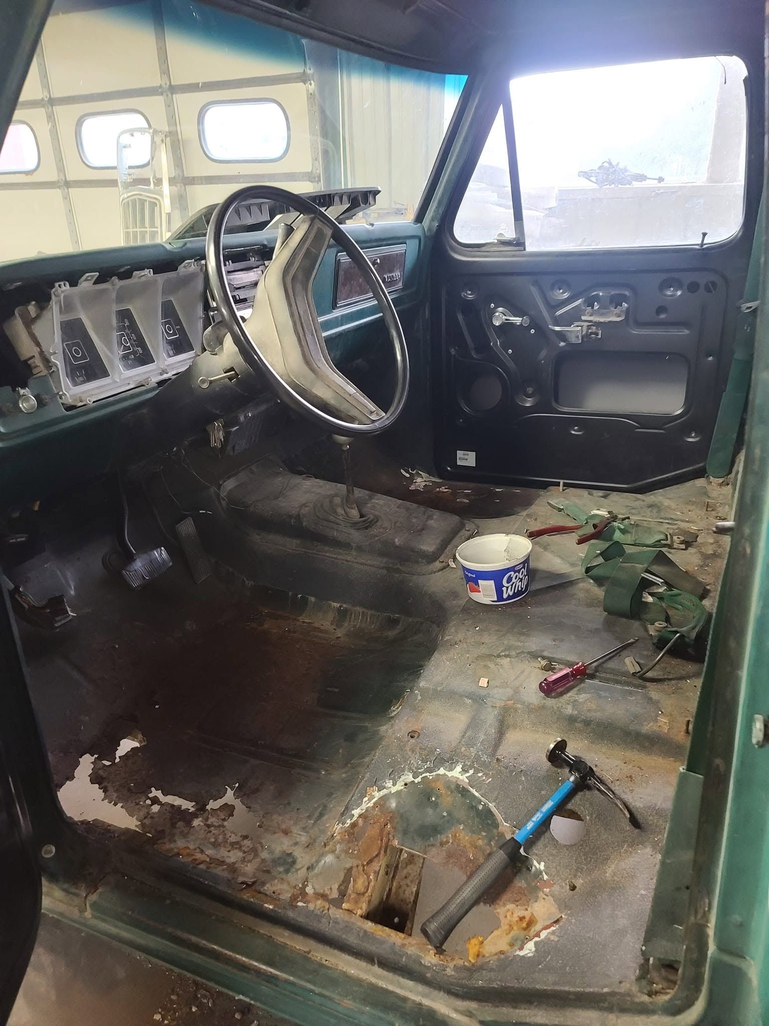 Rusty, stripped vehicle interior with torn floor, exposed metal, and a steering wheel by the driver’s seat