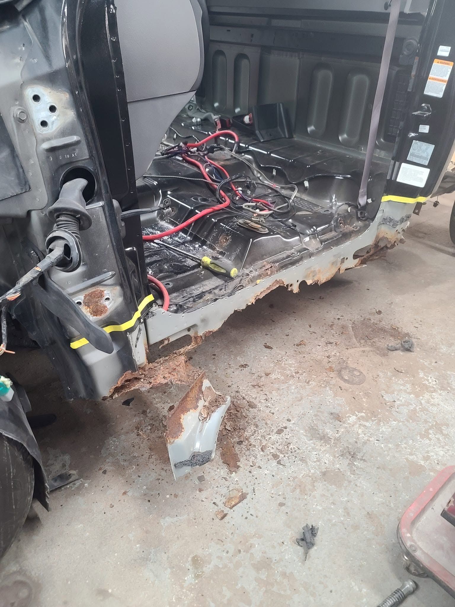 Damaged car interior with exposed wiring, rusted floor, and debris on the ground