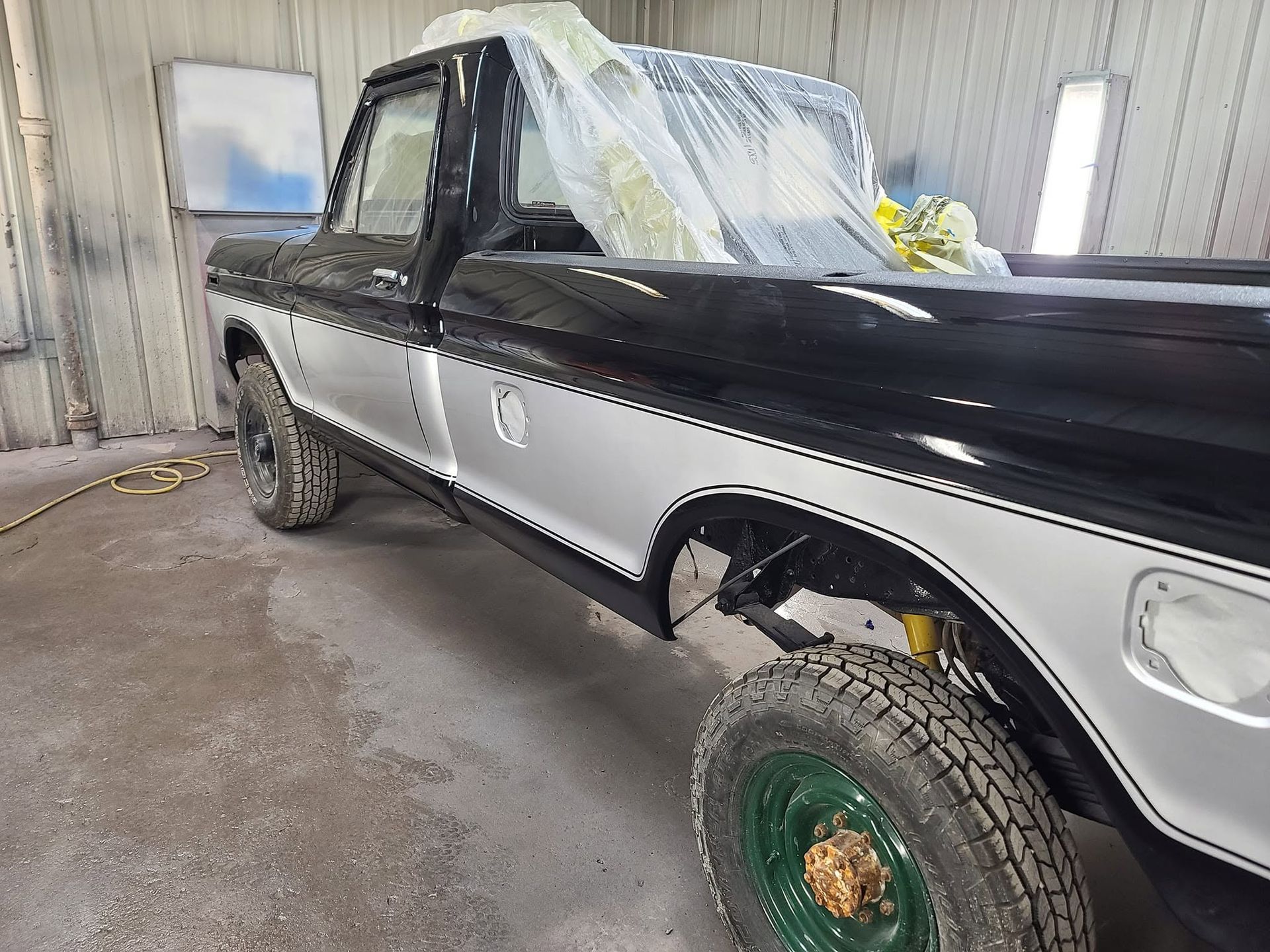 Black-and-silver pickup truck with windshield covered in plastic, parked inside a workshop with a missing front wheel.
