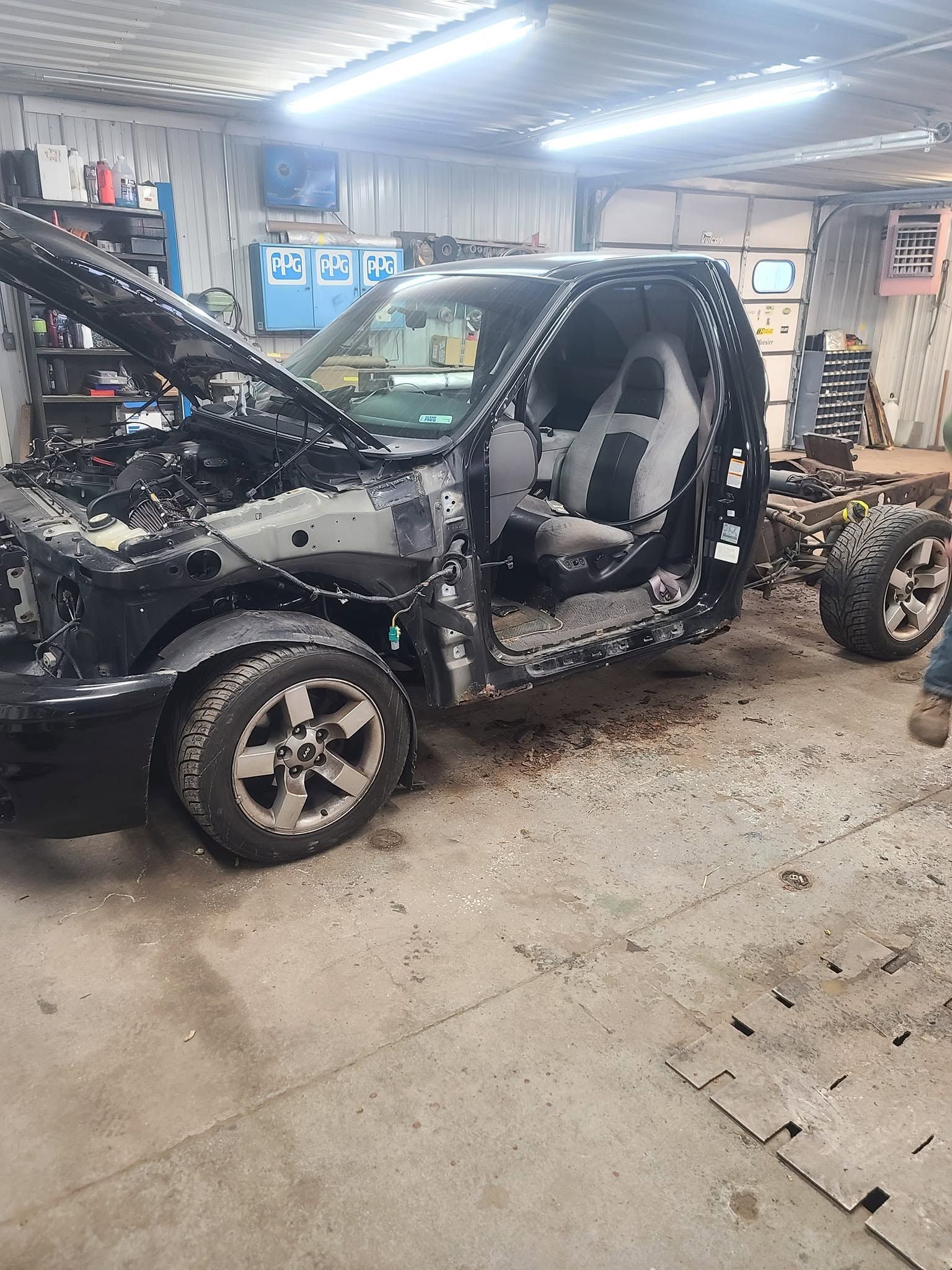 Black SUV with front damage in a repair shop, hood open and parts removed, under bright workshop lights