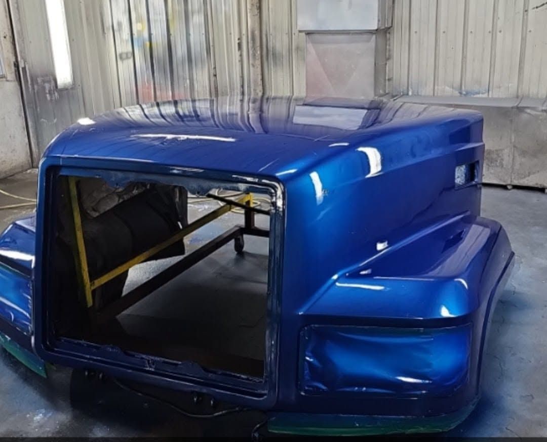 Blue car cab shell with large windshield opening, sitting in a workshop spray booth