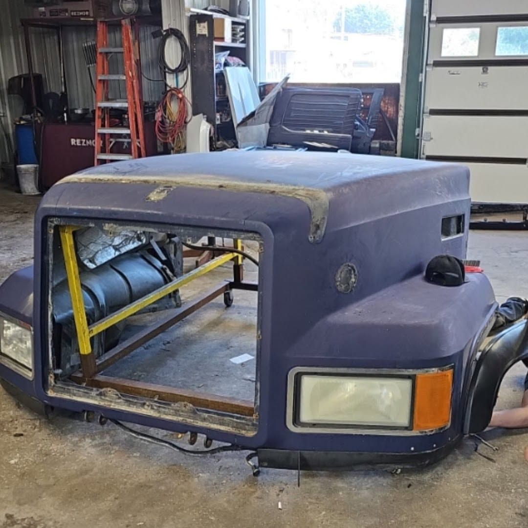 Dusty purple vehicle cab shell in a workshop, with exposed front frame and headlight opening.