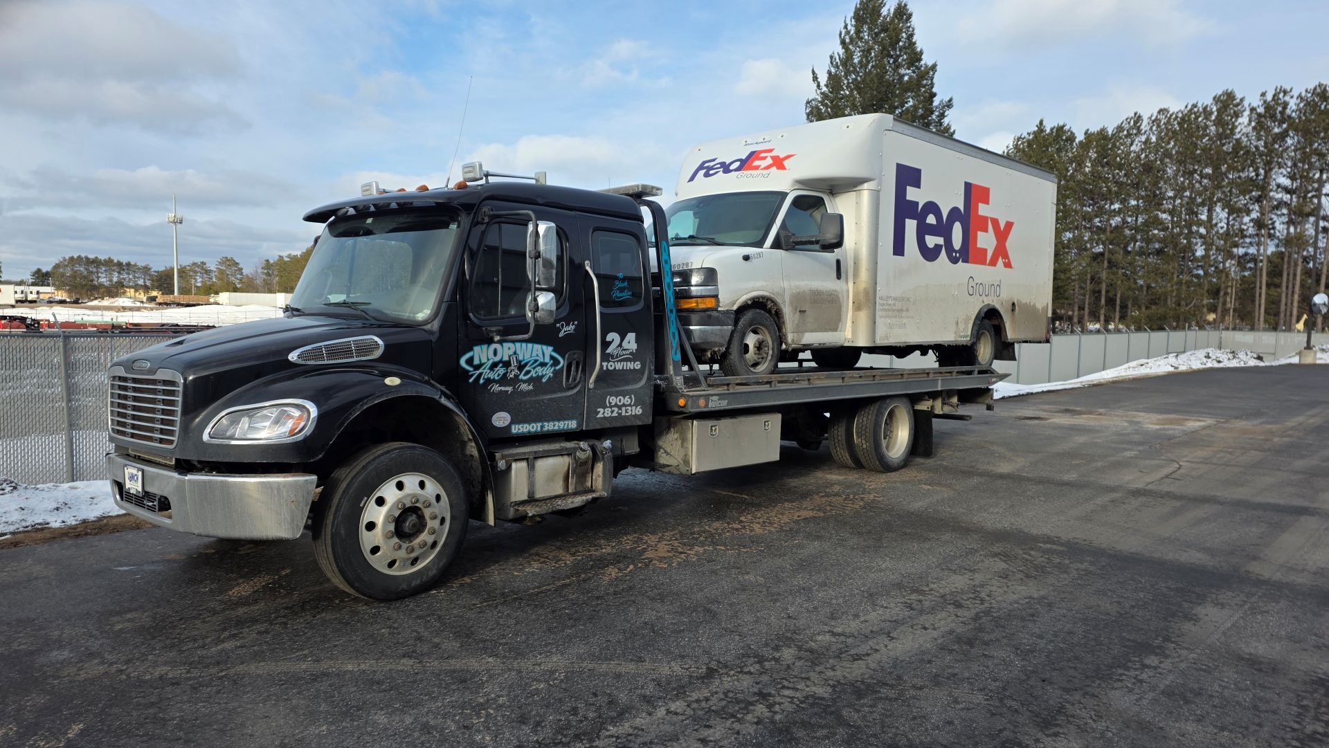 FedEx truck on a snowy road with a black tow truck in front, parked near a fenced area
