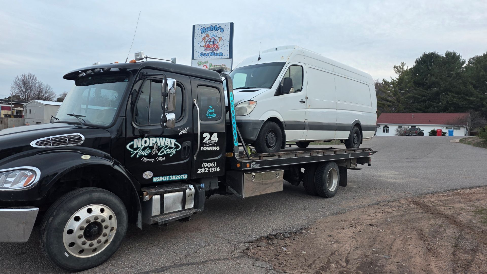 Black tow truck carrying a white van in a gravel lot under an overcast sky