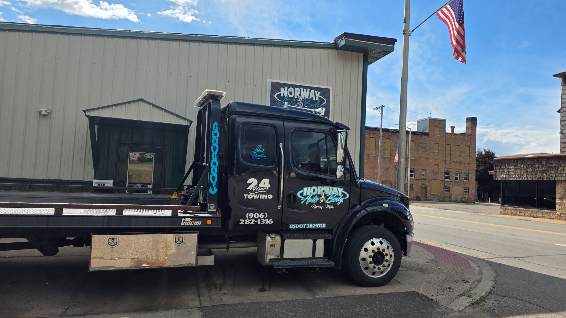 Black flatbed tow truck parked beside a gray industrial building with an American flag overhead.