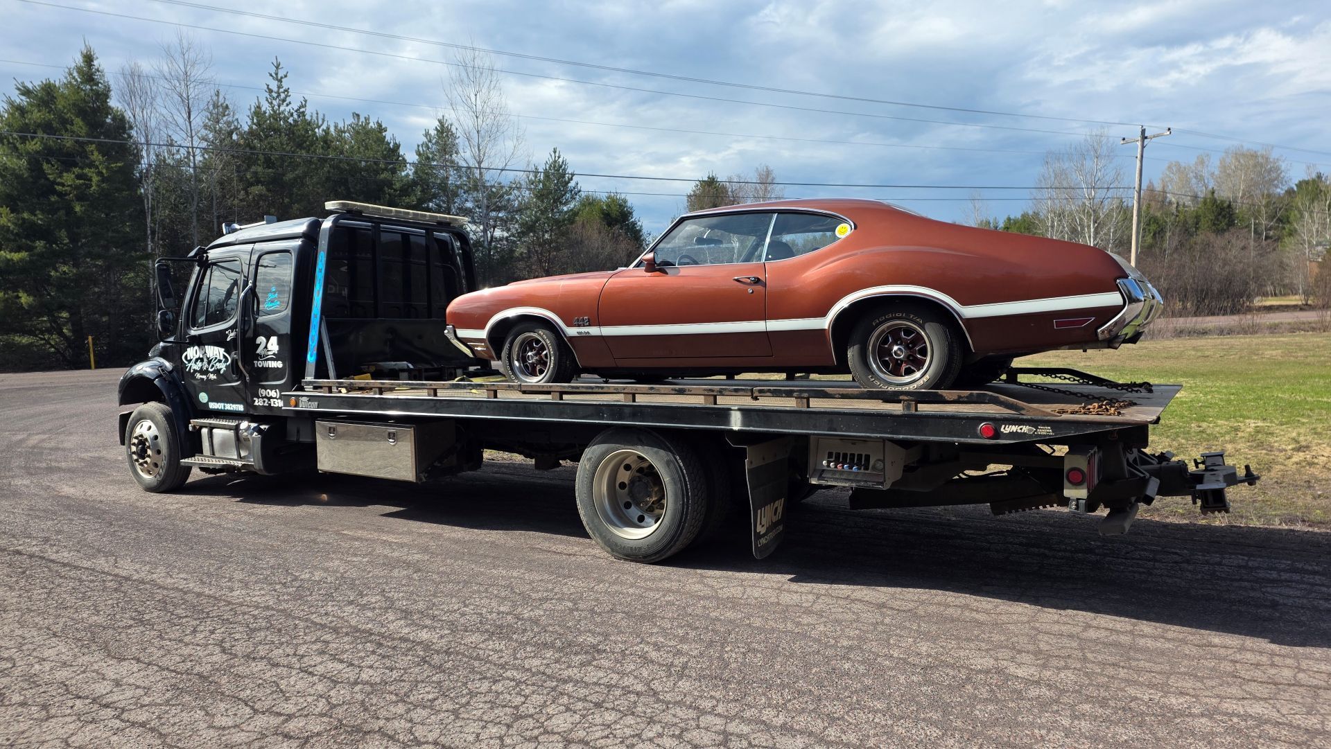 Flatbed tow truck carrying a rust-orange classic car on a road under cloudy skies