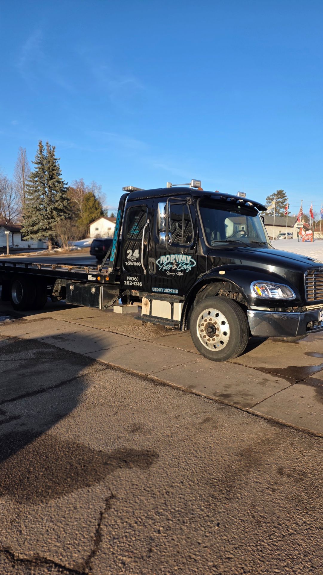 Black semi-truck parked on a sunny street beside a sidewalk and trees.