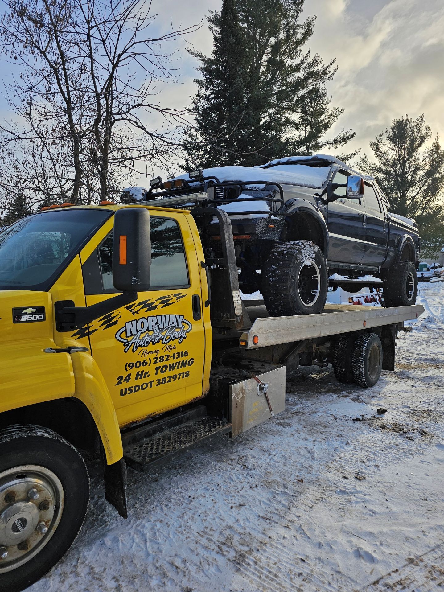 Yellow tow truck hauling a black SUV on a snowy roadside with bare trees