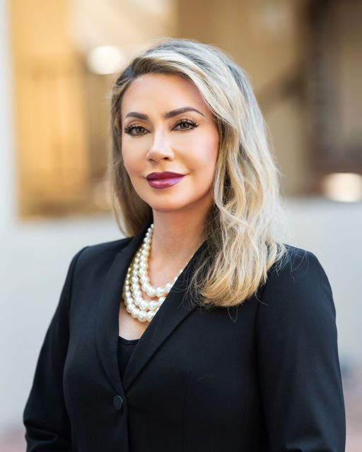Portrait of a woman in a black blazer and pearl necklace, standing outdoors with a blurred background