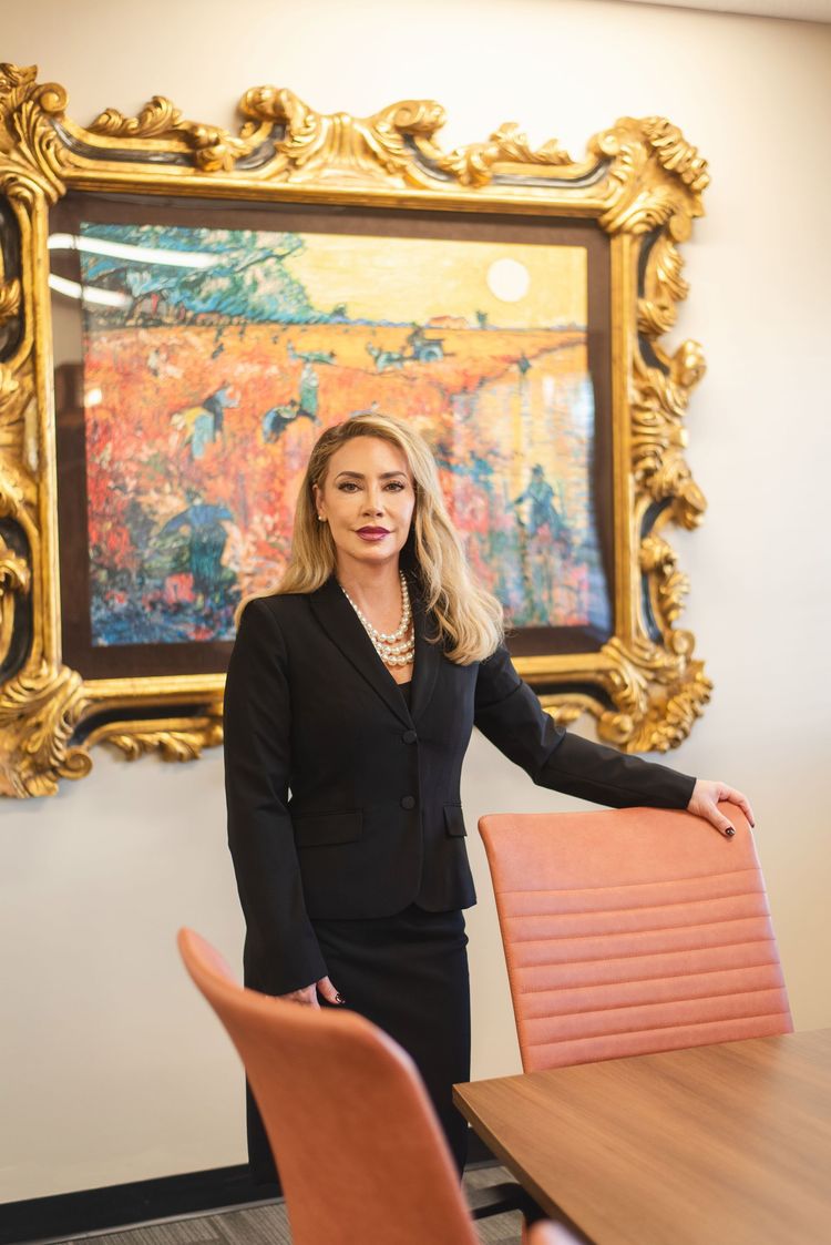 Woman in a black suit standing by a conference table in a stylish office with a gold-framed abstract painting.