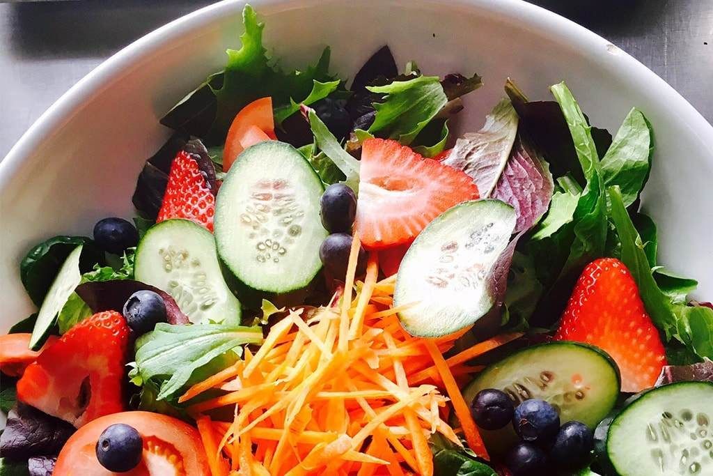 A salad with strawberries , cucumbers , carrots and blueberries on a white plate.