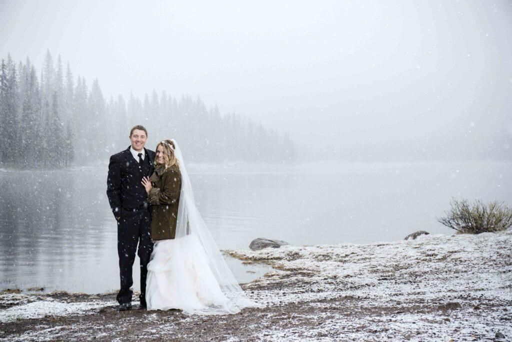 A bride and groom are posing for a picture in the snow near a lake.