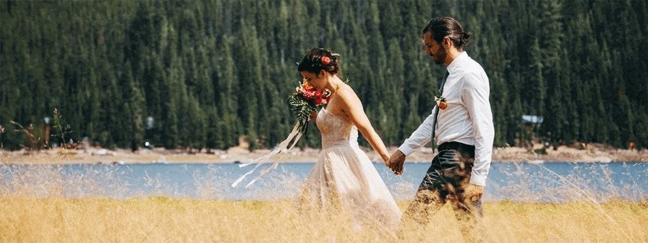 A bride and groom are walking through a field holding hands.
