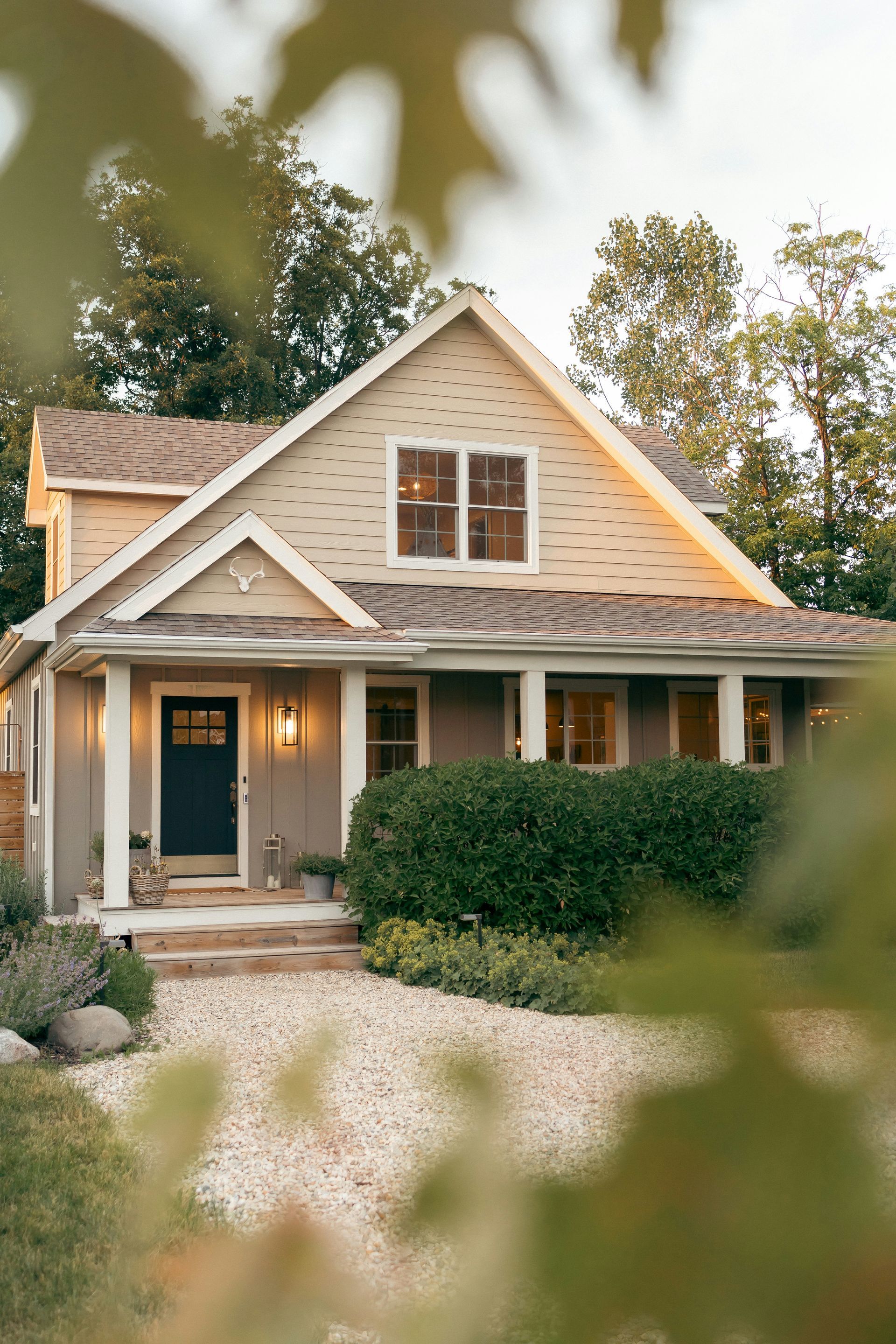 Charming beige cottage with a front porch, lit entryway, and trimmed hedge, viewed through soft foliage.
