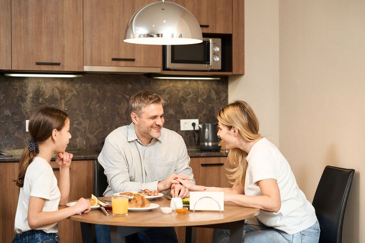 Three people talking at a dining table in a modern kitchen, sharing food and coffee