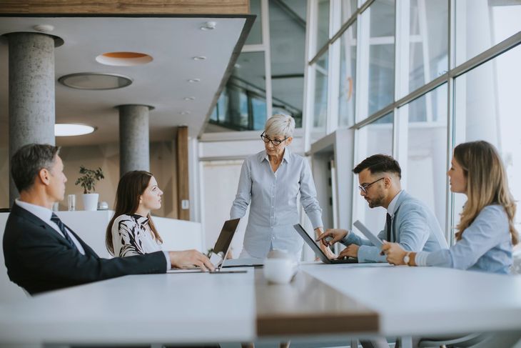 Team meeting in a bright office with five people around a table, one woman standing and speaking