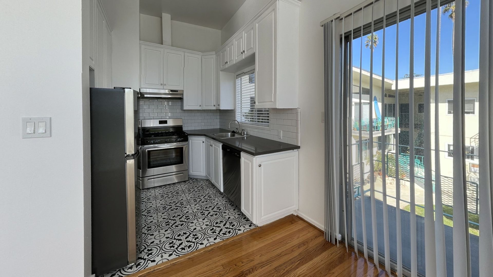 Small white kitchen with stainless steel appliances, dark countertops, patterned floor, and large window.