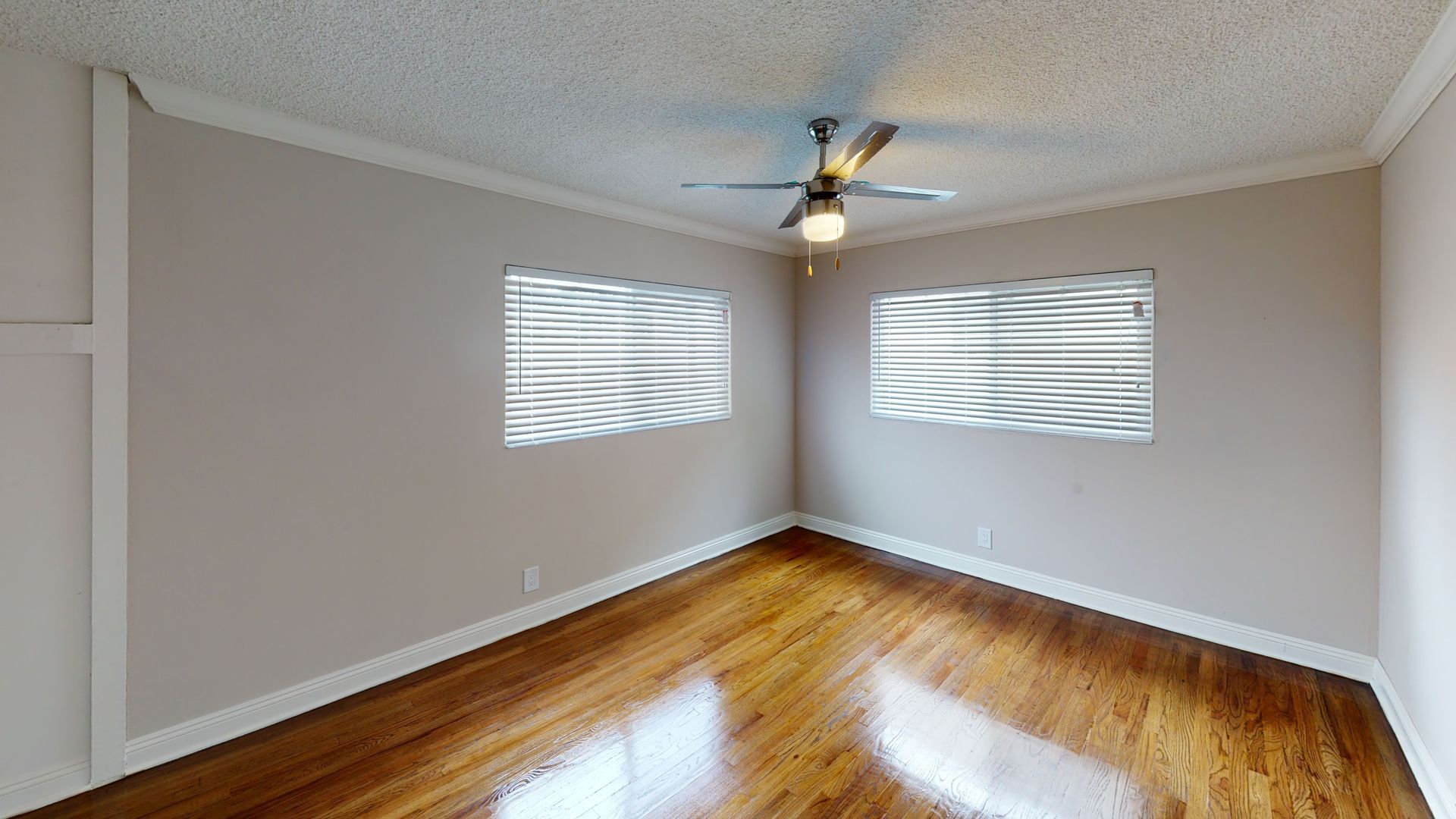 Empty room with hardwood floors, two windows, and a ceiling fan. Walls are painted light beige.