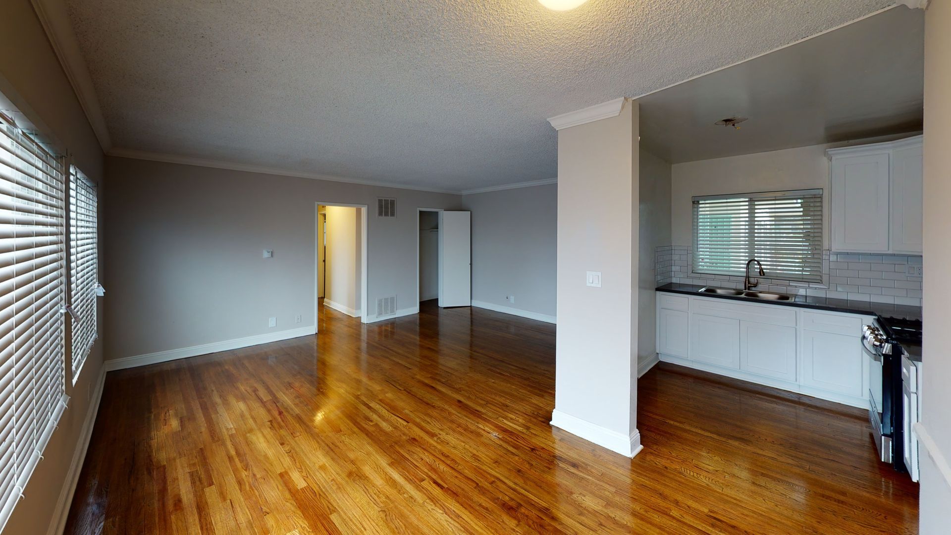Living room with hardwood floors, kitchen visible. Gray walls, white cabinets, and blinds.