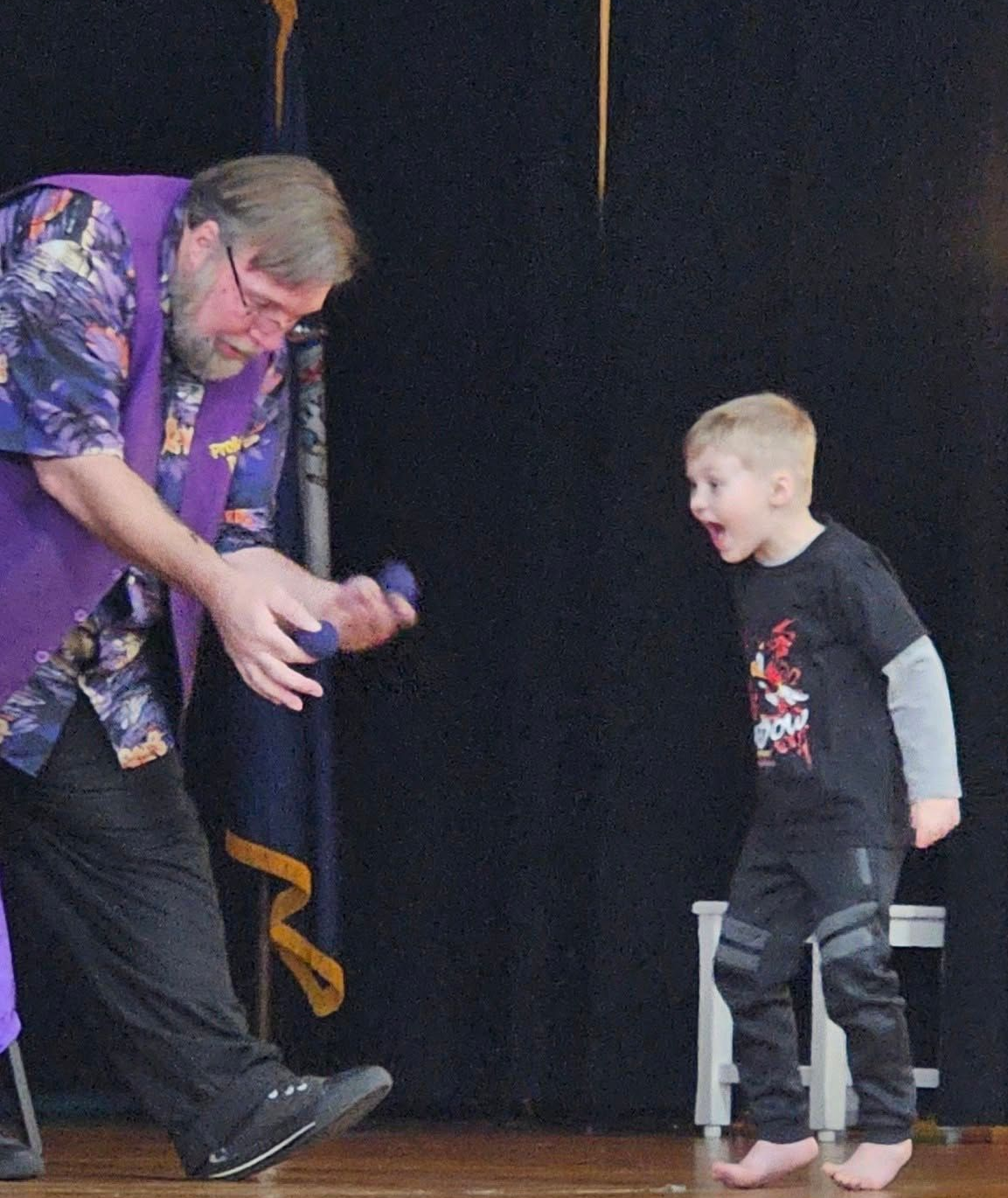 Magician in purple vest shows blue balls to excited child on stage. Black backdrop.