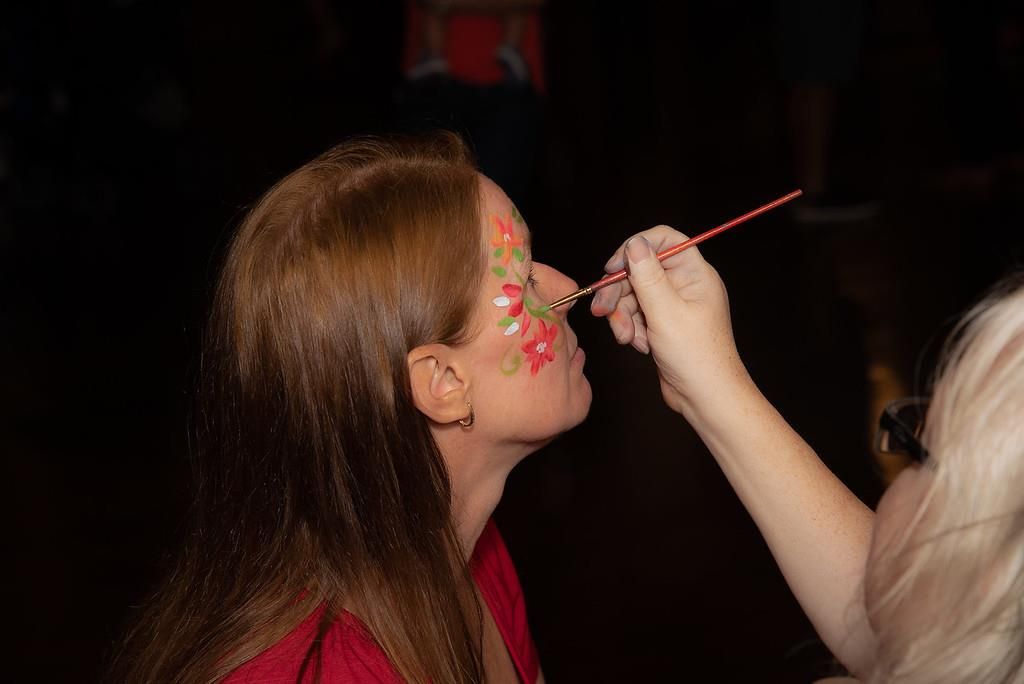 Woman getting face painted with red and green floral design. Artist's hand holds brush.