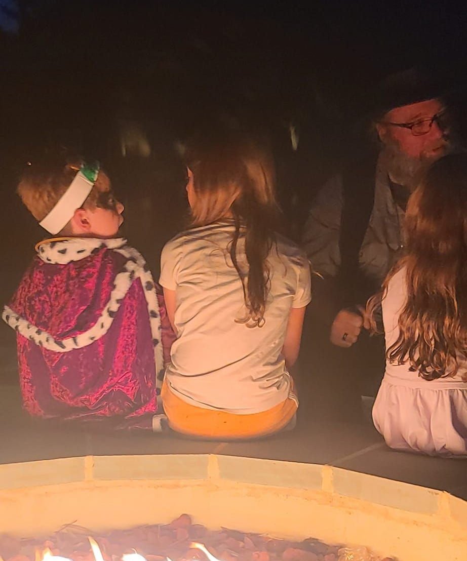 Children and an adult sit around a fire pit at night. One child is in a king's costume.