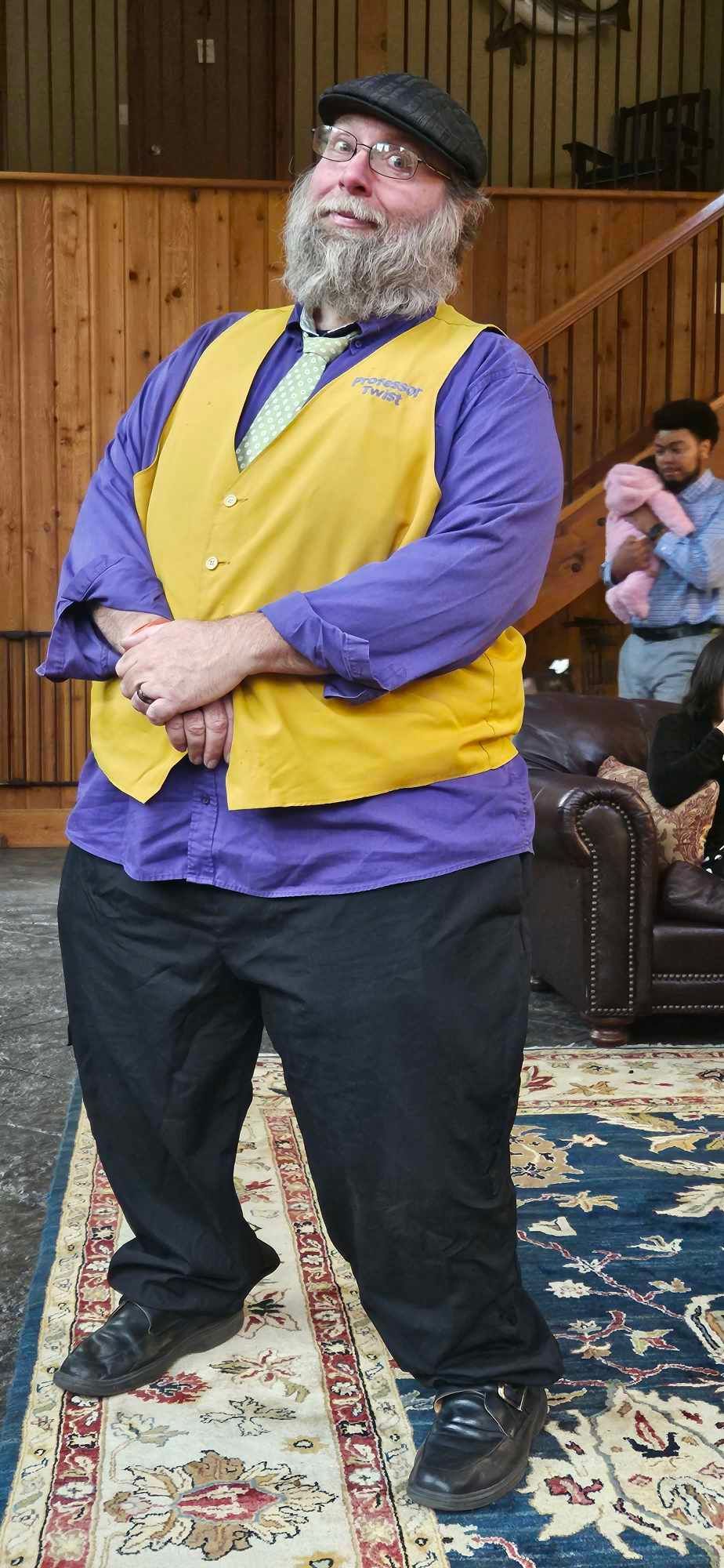 Man wearing a yellow vest and purple shirt standing indoors on a patterned rug, hands clasped
