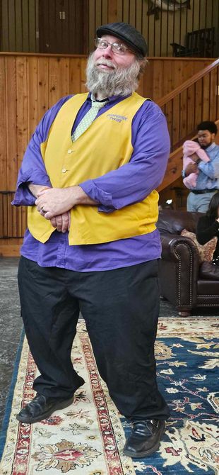 Man wearing a yellow vest and purple shirt standing indoors on a patterned rug, hands clasped